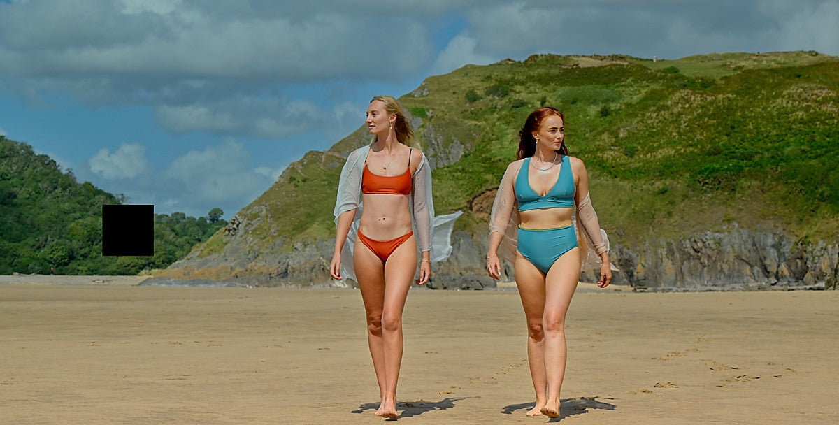 Two women in swimsuits walking on a beach with green hills in the background
