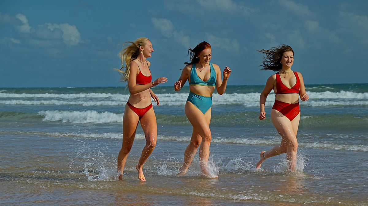 Three women in swimsuits running along a beach with ocean waves in the background