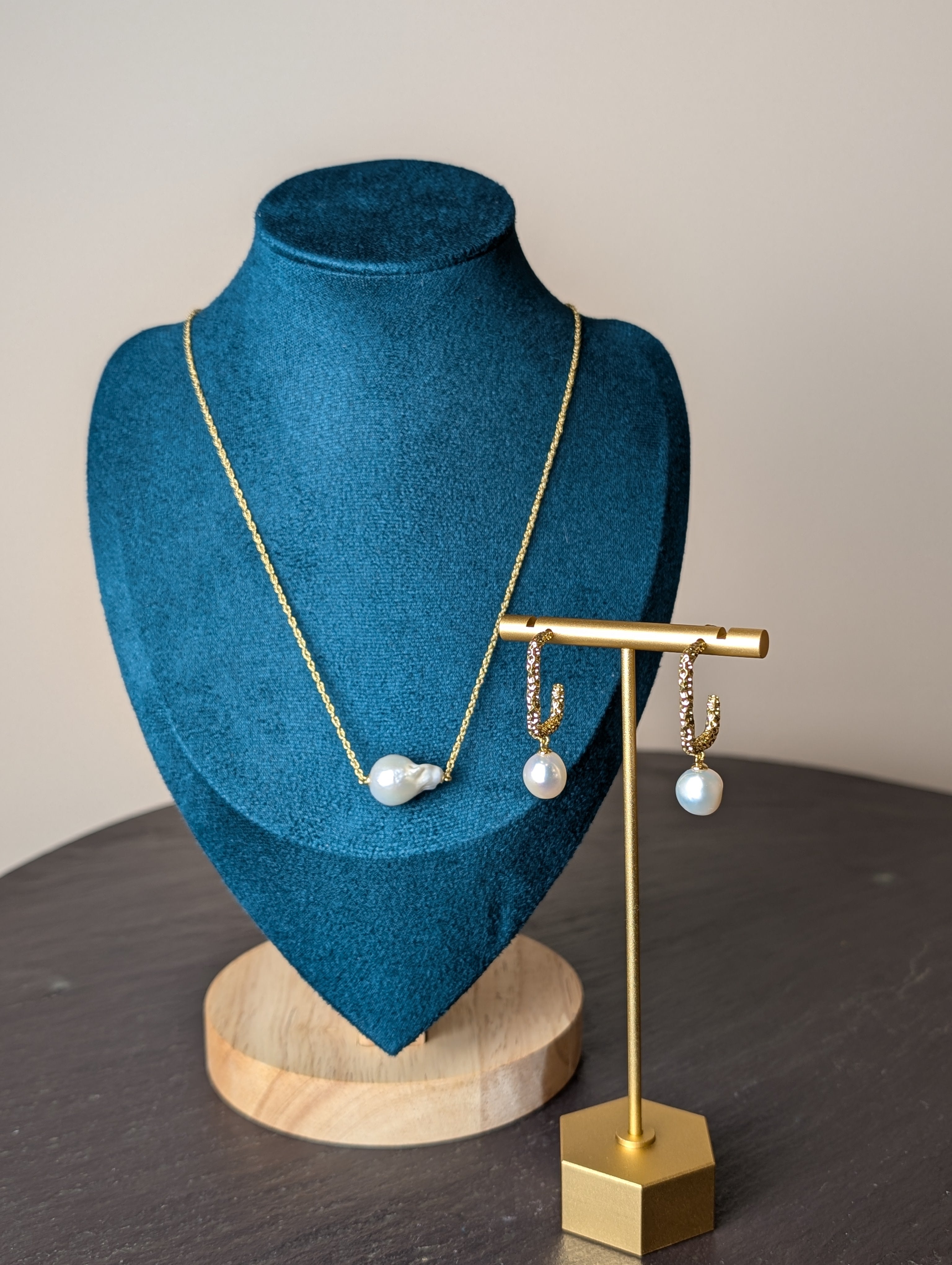 Nacre Duo Set by Nudie – studio photo showing the freshwater pearl necklace on a green and wooden mannequin, with matching earrings displayed on a gold stand atop a slate table.