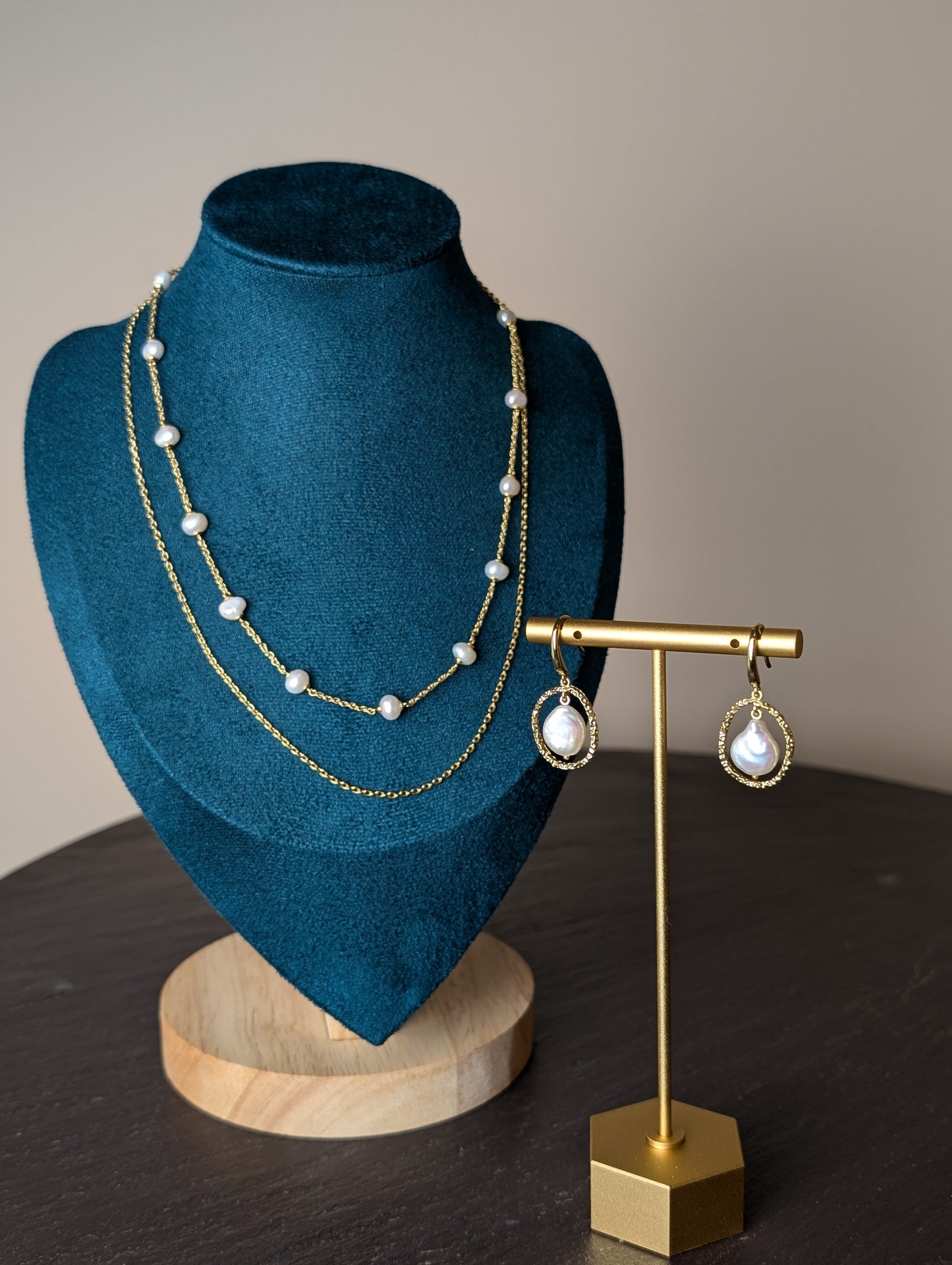 Maris Duo Set by Nudie – studio photo showing freshwater pearls on a fine gold chain, layered with a second twisted strand necklace on a green and wooden mannequin, with matching earrings on a gold stand atop a slate table.