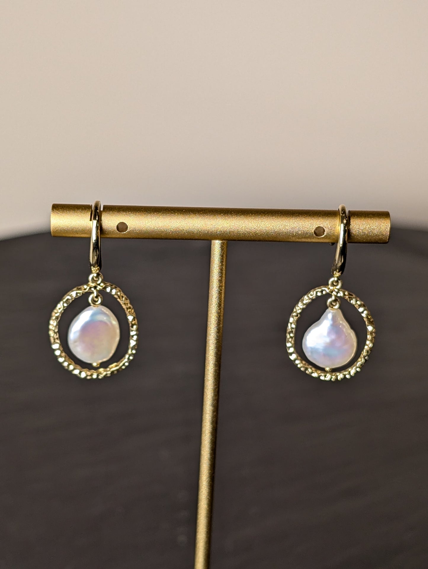 Maris Earrings by Nudie – close-up studio photo showing teardrop freshwater pearls suspended inside hammered gold hoops, displayed on a gold earring stand.