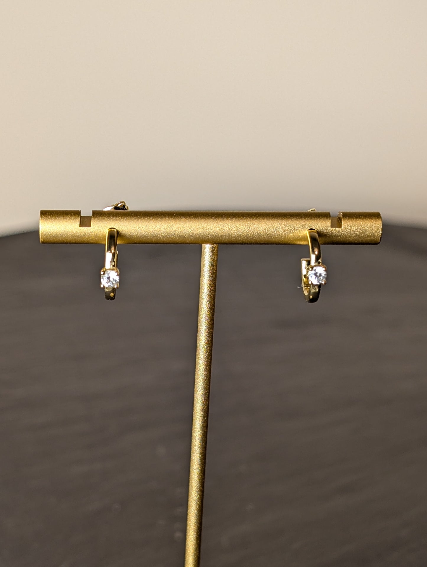 Avalon Earrings by Nudie – close-up studio photo showing hammered gold hoops on a gold earring stand, with a softly blurred background and slate table beneath.