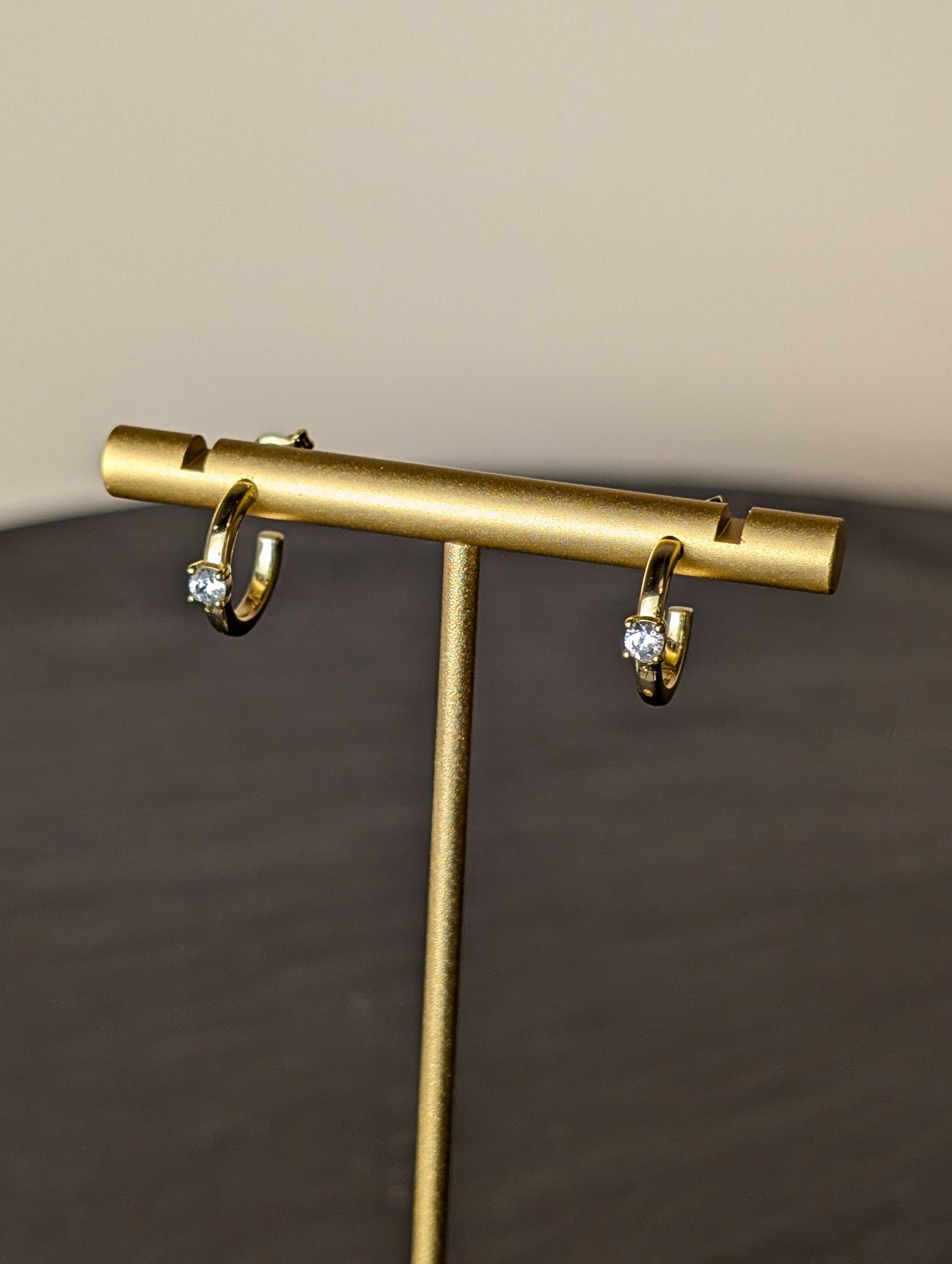 Avalon Earrings by Nudie – close-up studio photo showing hammered gold hoops on a gold earring stand, with a softly blurred background and slate table beneath.