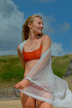 Woman in an orange bikini top and white cover-up standing on a beach with greenery and blue sky in the background.