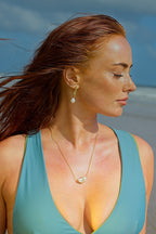 Woman in a teal swimsuit on a beach with a necklace and earrings.