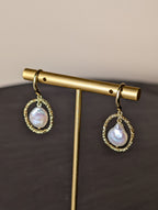 Maris Earrings by Nudie – close-up studio photo showing teardrop freshwater pearls suspended inside hammered gold hoops, displayed on a gold earring stand.