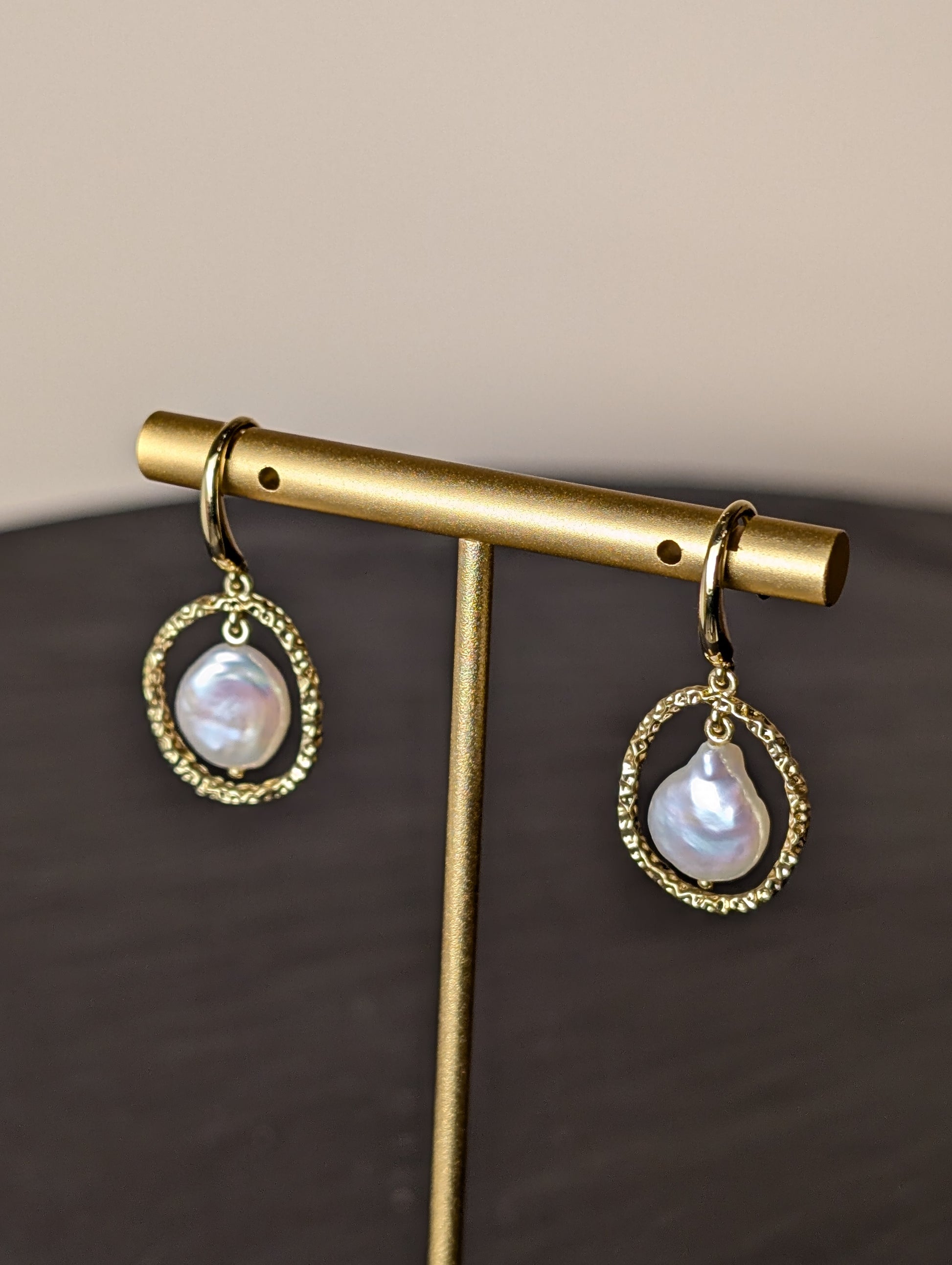 Maris Earrings by Nudie – close-up studio photo showing teardrop freshwater pearls suspended inside hammered gold hoops, displayed on a gold earring stand.