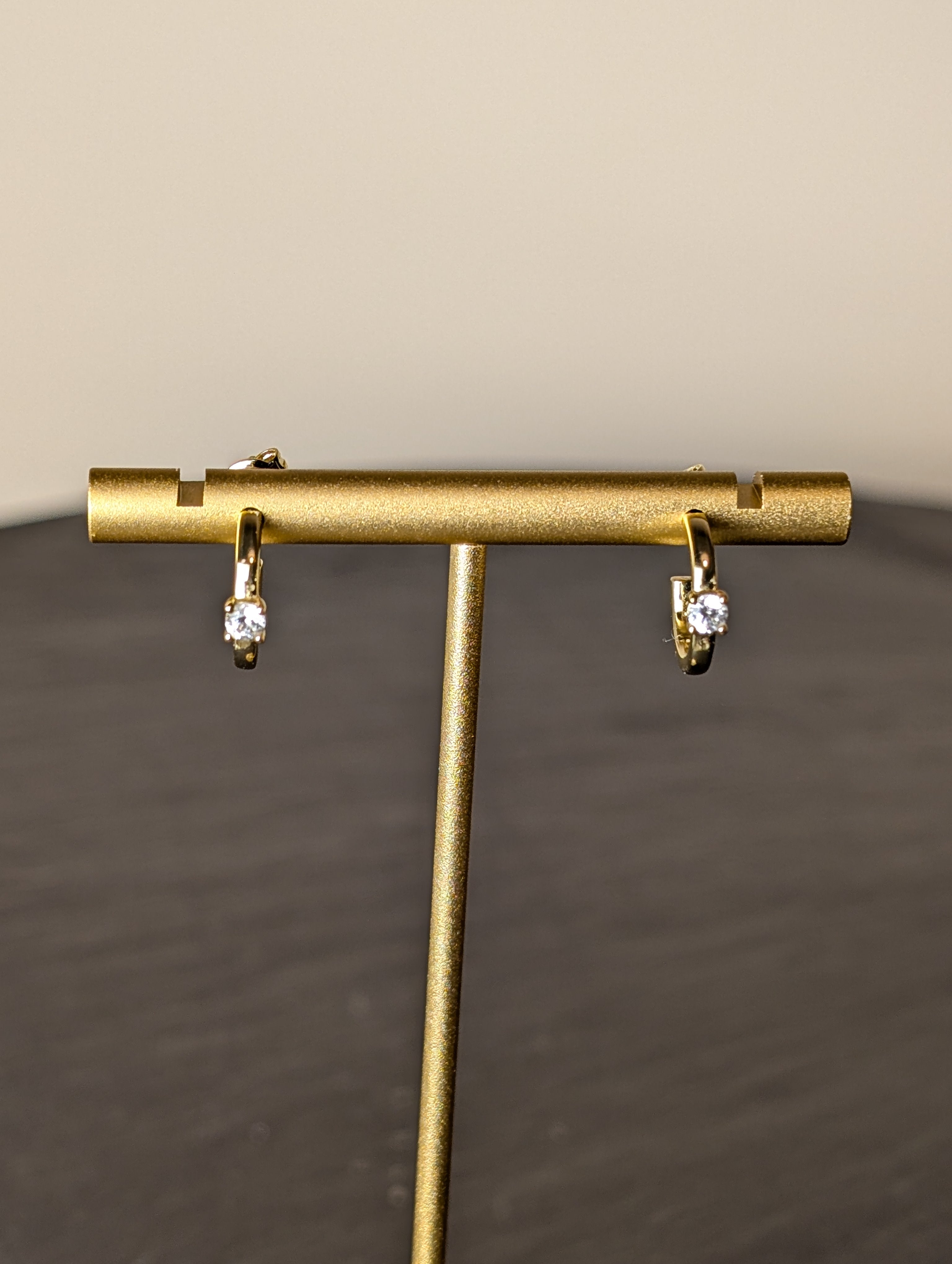 Avalon Earrings by Nudie – close-up studio photo showing hammered gold hoops on a gold earring stand, with a softly blurred background and slate table beneath.