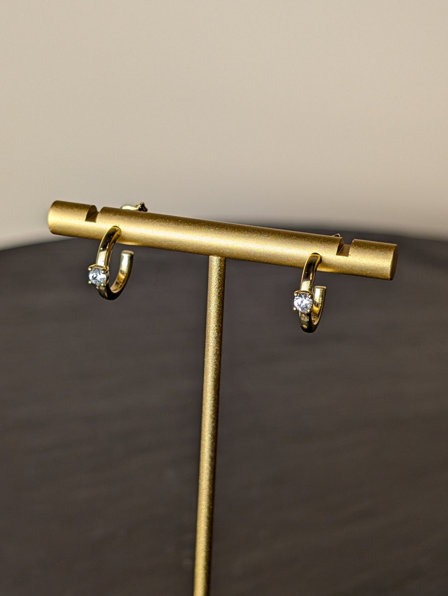 Avalon Earrings by Nudie – close-up studio photo showing hammered gold hoops on a gold earring stand, with a softly blurred background and slate table beneath.