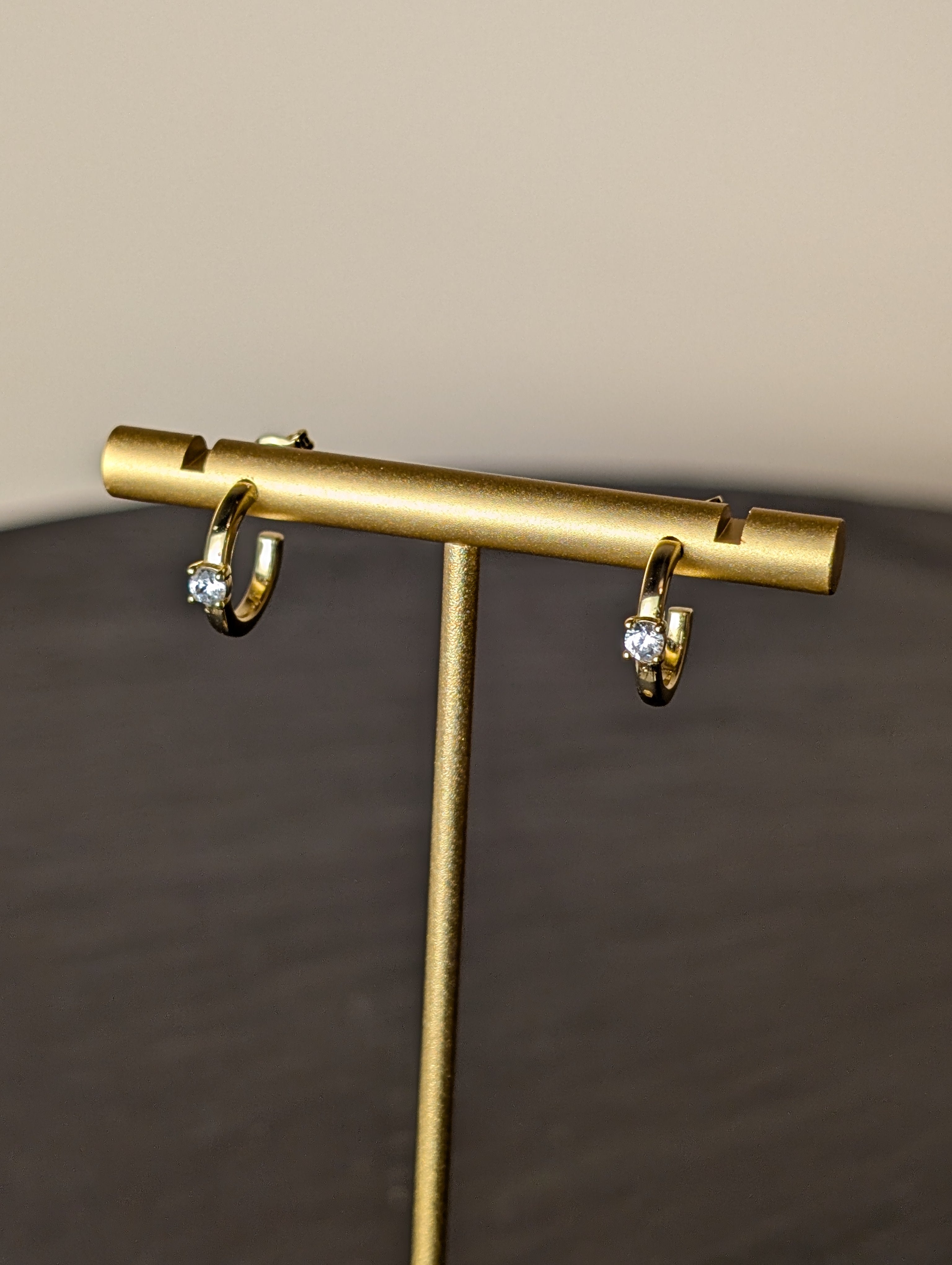 Avalon Earrings by Nudie – close-up studio photo showing hammered gold hoops on a gold earring stand, with a softly blurred background and slate table beneath.