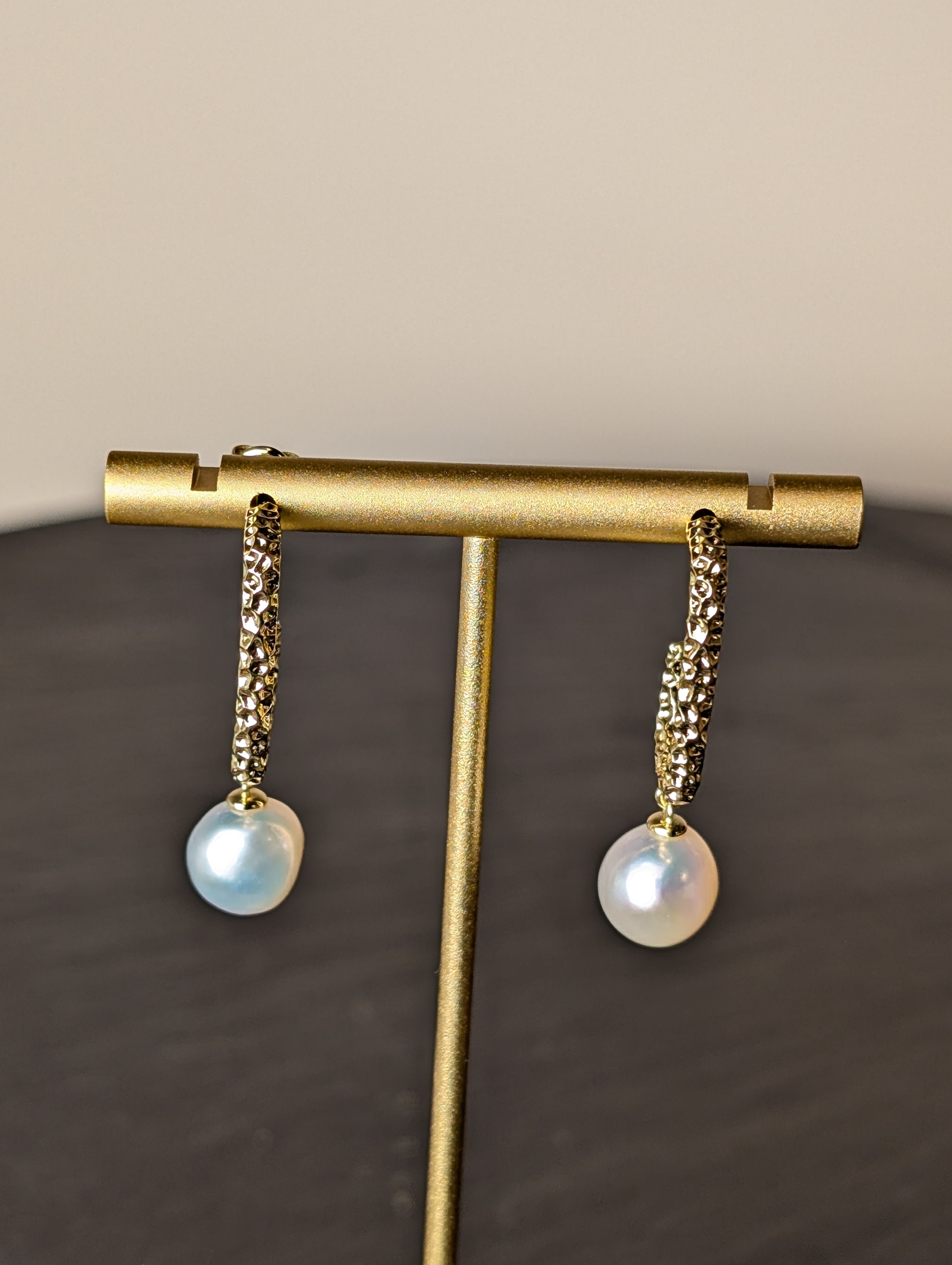 Nacre Earrings by Nudie – close-up studio photo showing a side-on view of the hammered gold earring with irregular freshwater pearl, displayed on a gold stand.