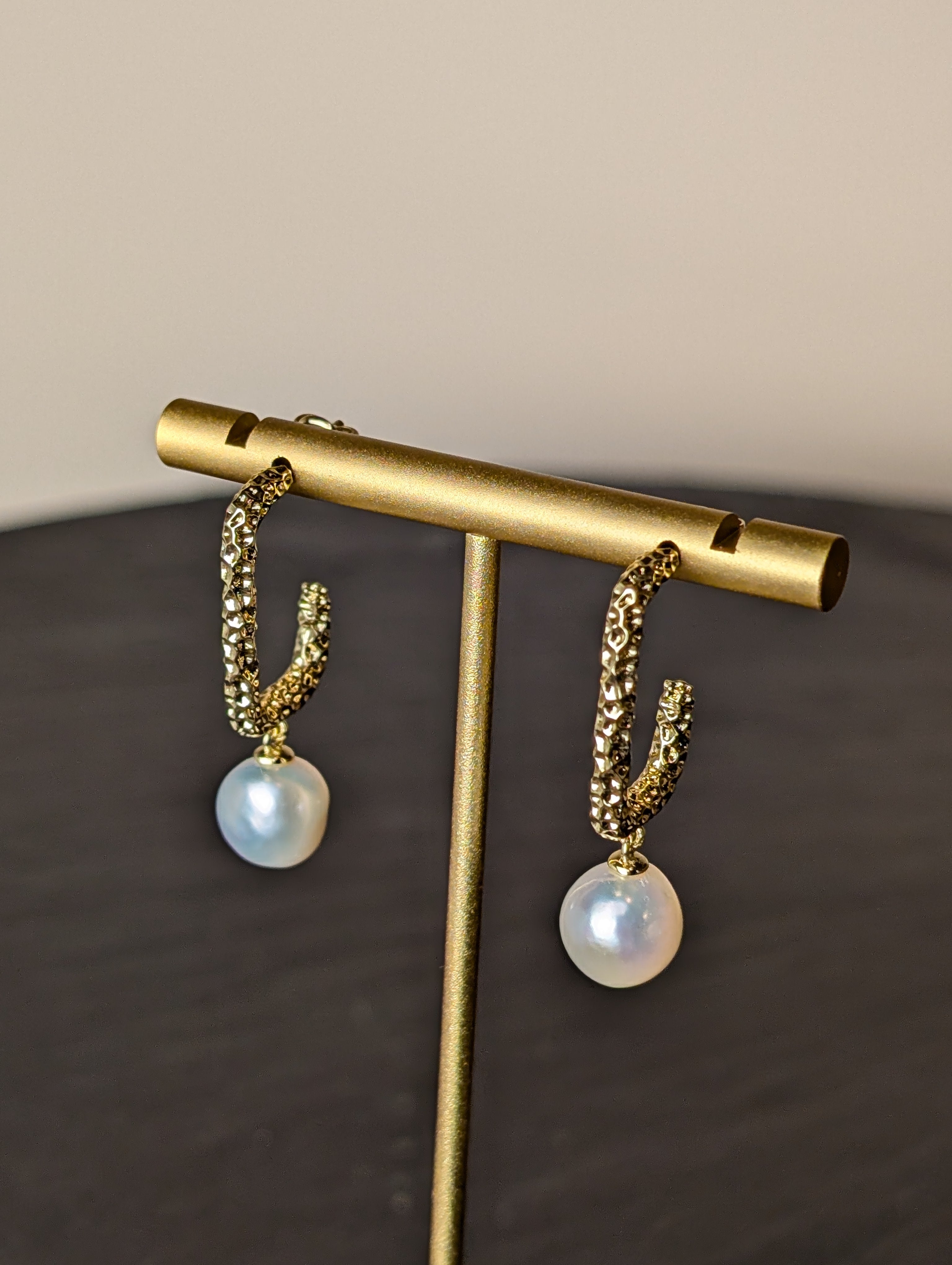 Nacre Earrings by Nudie – close-up studio photo showing a side-on view of the hammered gold earring with irregular freshwater pearl, displayed on a gold stand.